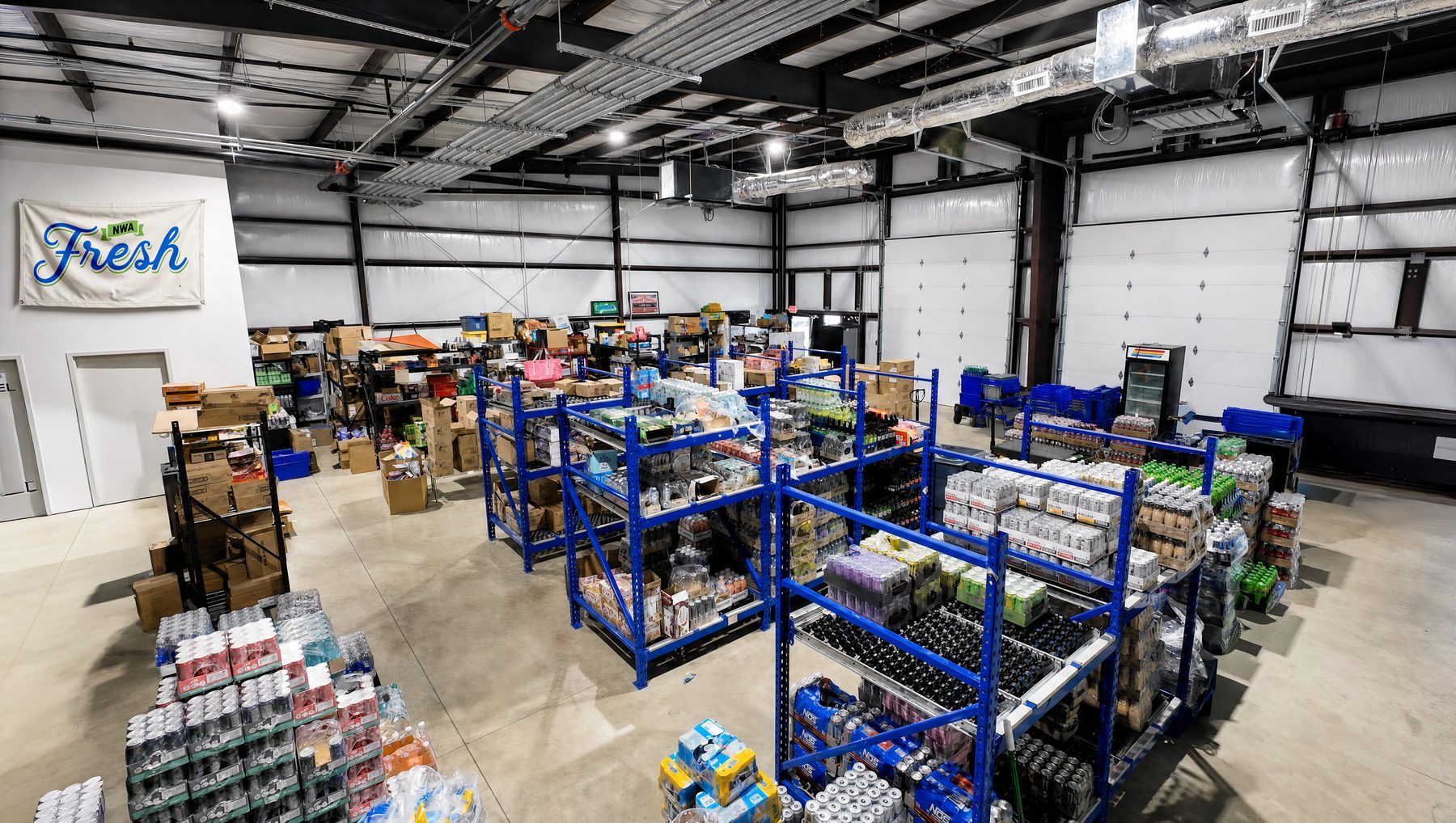 The NWA Fresh warehouse — low blue shelving in island rows, hand-stacked cases, roll-up doors